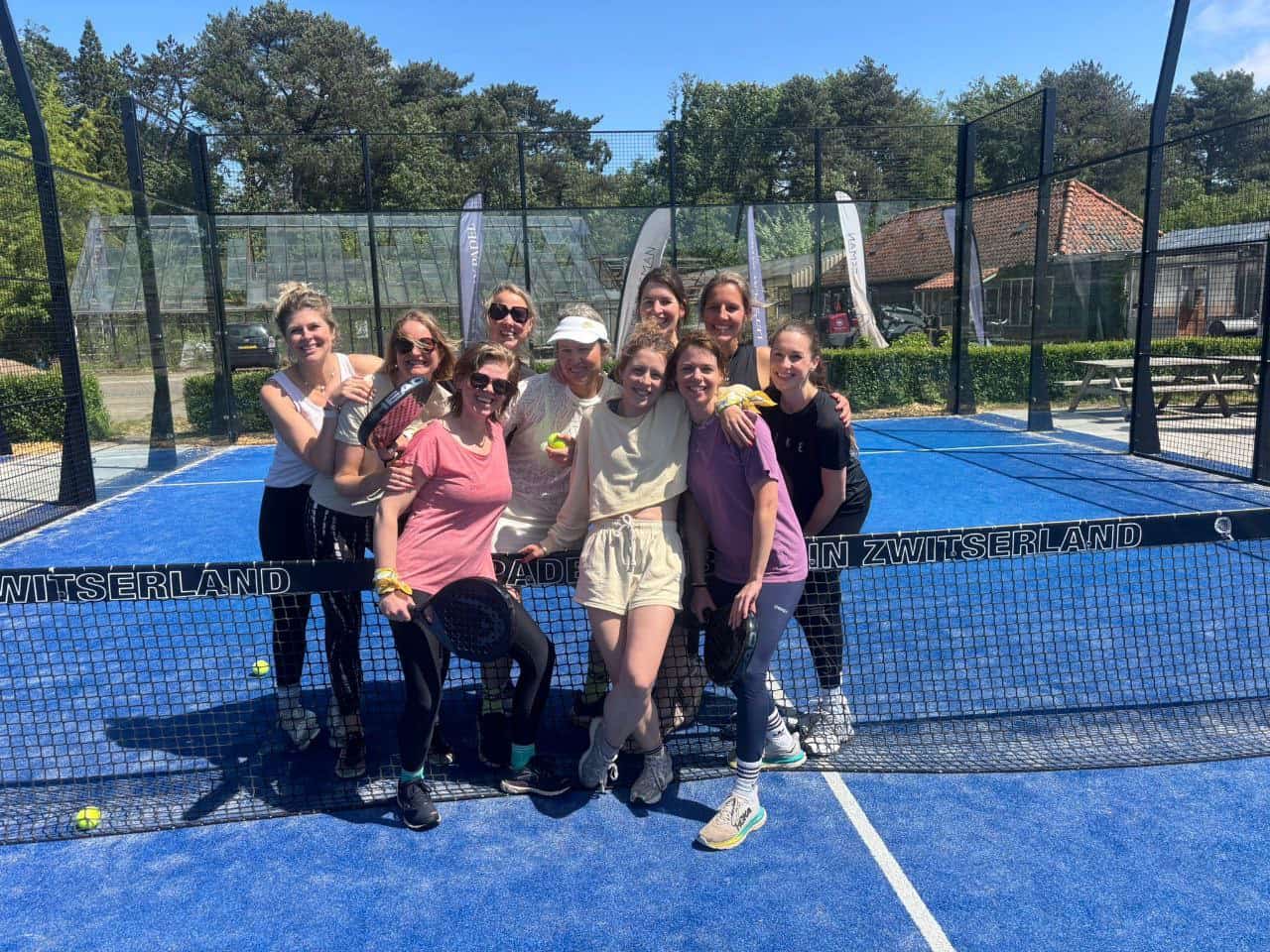 Groep enthousiaste vrouwen op padelbaan tijdens NXT Level Padelacademy, genietend van sport en gezelligheid in een zonnige buitenomgeving.