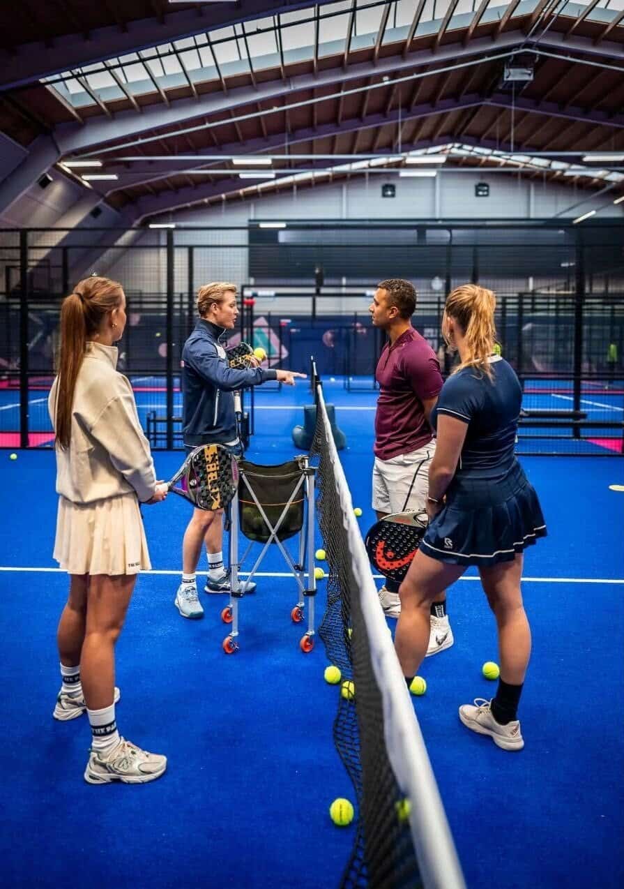 Professionele padeltraining met coach op indoor padelbaan, jonge sporters leren techniek en tactiek, NXT Level Padelacademy, sportieve ambiance, tennisrackets en ballen aanwezig, maximale prestatietraining.